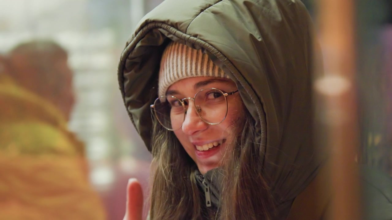 Young woman in hood gives thumbsup at nighttime bus shelter, beanie and glasses, soft bokeh lights, candid smile, grainy film texture, warm jacket, urban transit scene, supportive gesture, friendly