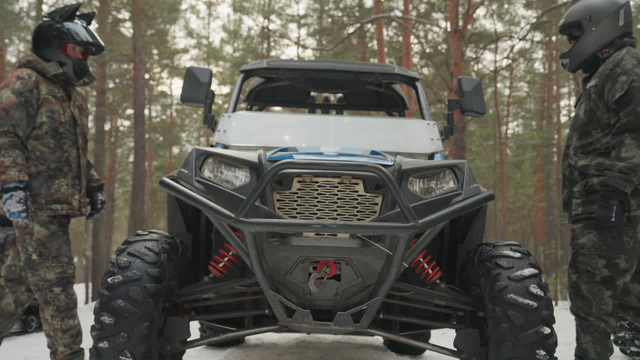 Primo piano frontale di un ATV con l'equipaggio in una pineta innevata. Il paraurti robusto, la griglia, i pneumatici e le sospensioni dominano l'inquadratura. Due piloti si scambiano segnali durante il controllo di sicurezza pre-corsa, mentre i meccanici ispezionano il verricello.