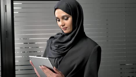 una joven hermosa con hijab negro usa una tableta, mira fotos. mujeres árabes en la oficina. 60 fps