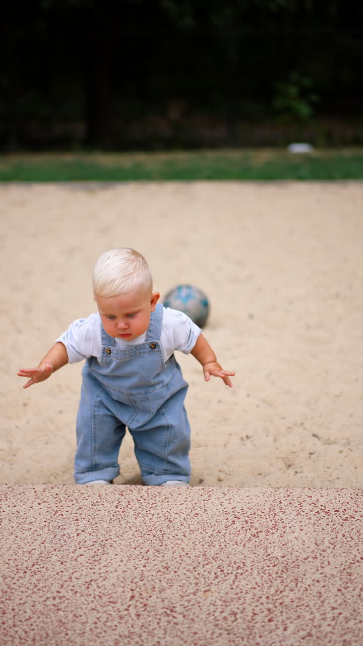 Adorable blond kid in jeans romper walks by the sand. Toddler boy leans to pick some sand. Vertical video.