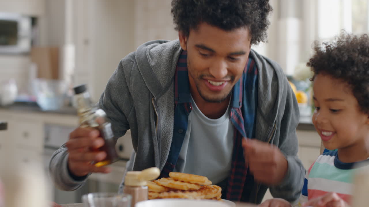 bambino felice che mangia waffle fresche con la famiglia a colazione padre che versa miele che prepara deliziosi dolcetti fatti in casa per il figlio in cucina a casa 4k