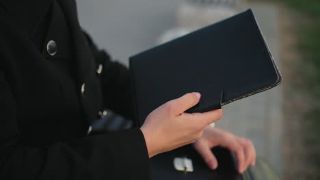 close up of individual hand in black clothing opening briefcase, searching through documents, retrieves black book, background features interlocked path, stone structure, greenery in outdoor setting