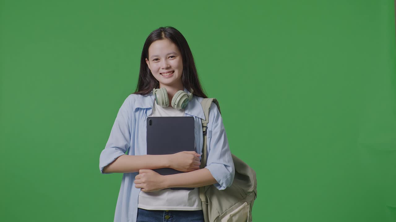Asian Teen Girl Student With A Backpack Holding Tablet And Smiling To Camera While Standing In The Green Screen Background Studio