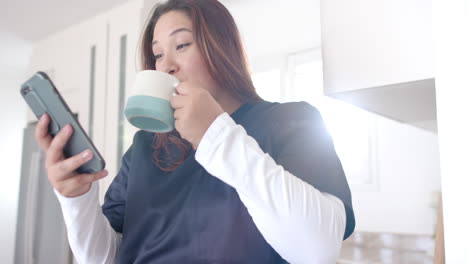 happy plus size biracial female doctor having coffee using smartphone at home, slow motion