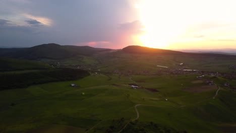 Aerial-pan-of-a-sunset-over-wide-fields-and-green-meadows-near-a-town,-Transylvania,-Romania