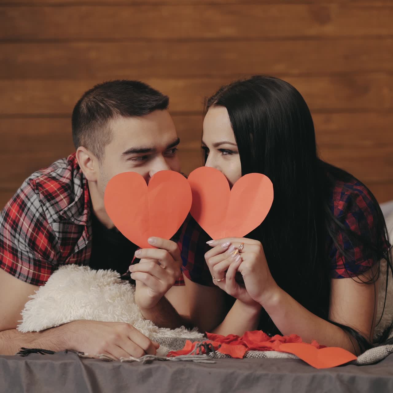 Adult man and woman in plaid shirts are lying on the bed and peeking out from under red paper hearts in their hands in the studio. Valentine's Day. Love story
