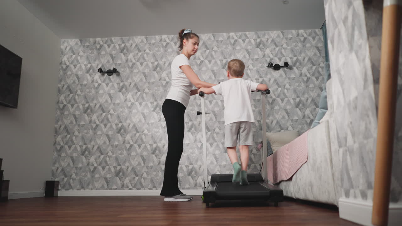 Focused mother gently wipes sweat from young boy face as he exercises on treadmill, showing care and encouragement during home workout session in cozy living room with visible TV and wooden seat leg