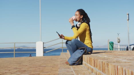 African-american-woman-sitting-using-smartphone-drinking-coffee-on-promenade-by-the-sea