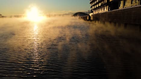 Vuelo-Bajo-Aéreo-Sobre-Aguas-Tranquilas-Junto-A-Apartamentos-Junto-Al-Agua-Con-Niebla-Flotante,-Inclínelo-Hacia-Arriba-Para-Revelar-La-Puesta-De-Sol-De-La-Hora-Dorada-Cerca-De-Bjorvika,