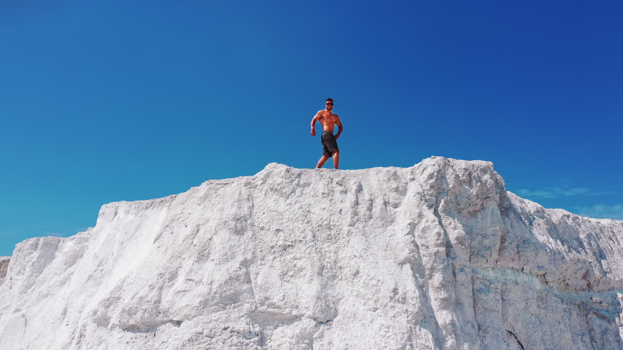 Drone view on the high white mountain and muscular man on top of it in summer. Topless young sportsman showing his trained body while standing on the top of the hill under blue sky.