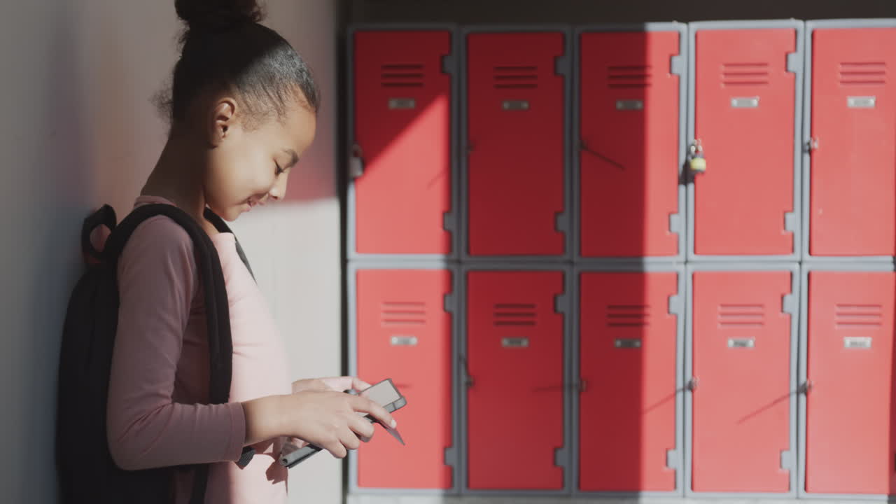 Using smartphone, African American girl standing by lockers in school hallway, smiling, copy space