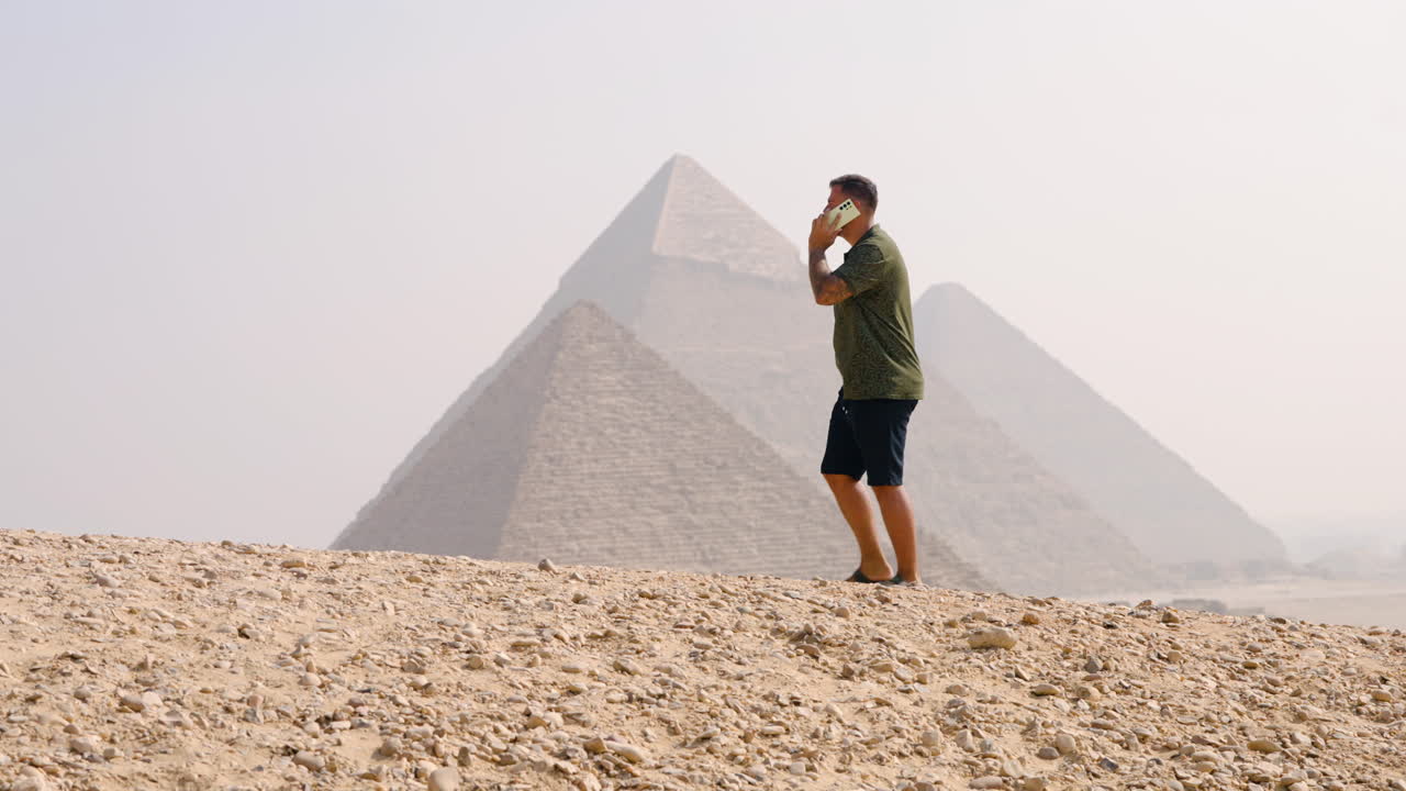 A Man Speaking on the Phone While on the Giza Plateau, With Views of the Pyramids in Cairo, Egypt - Handheld Shot