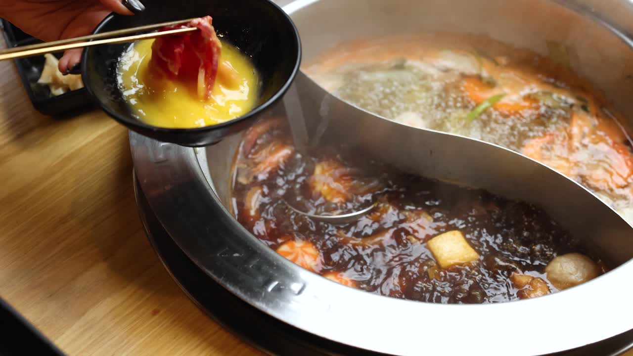 Chopsticks dip raw beef into beaten egg beside steaming dual-broth hot pot at restaurant table