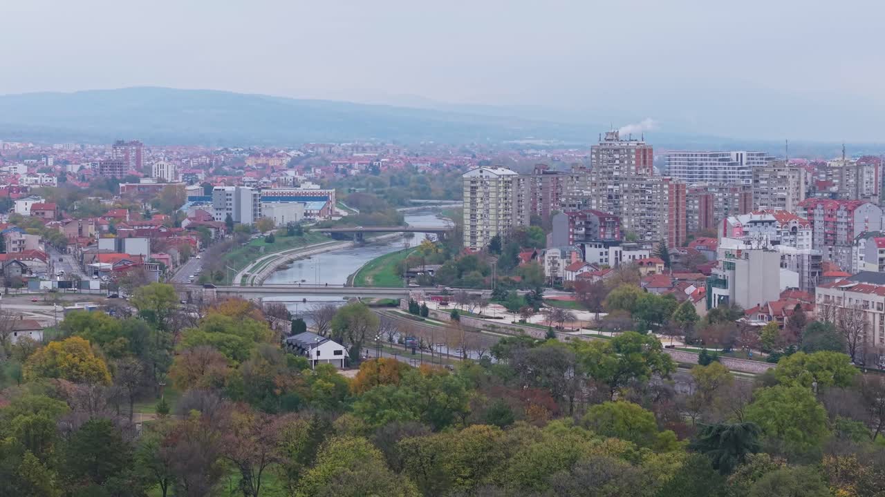 Un drone si alza da dietro gli alberi, rivelando gradualmente la tentacolare città di Niš, in Serbia, fondendo il verde con i tetti e le strade urbane in una vista aerea cinematografica