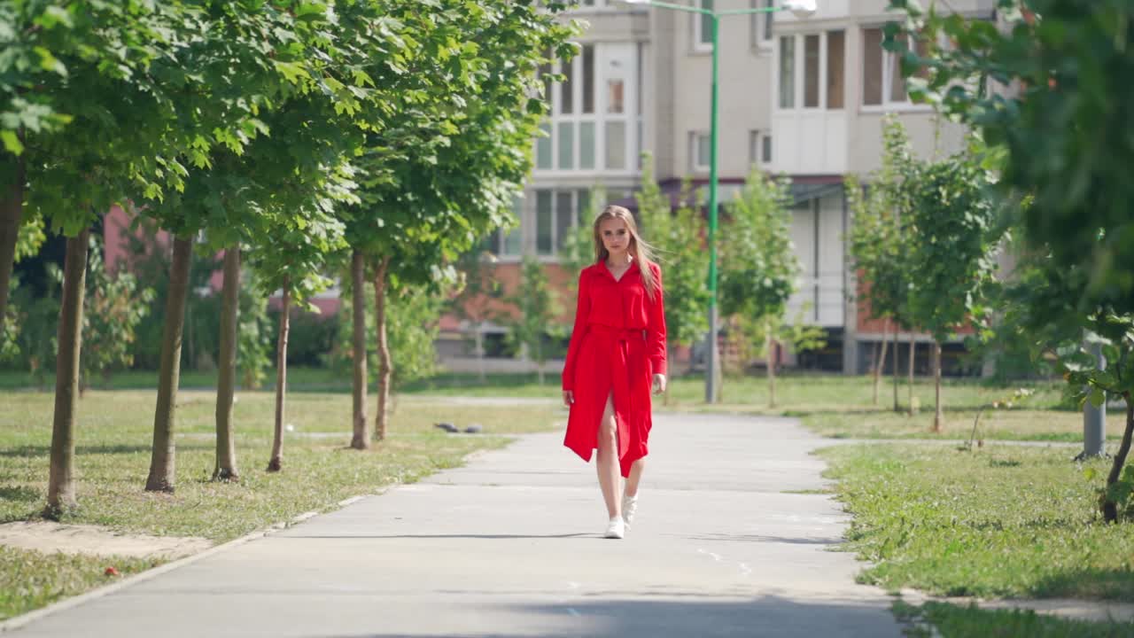 Full-length portrait of a pretty model. Young woman in red dress going forward in the city street. Attractive girl outdoors. Slow motion.