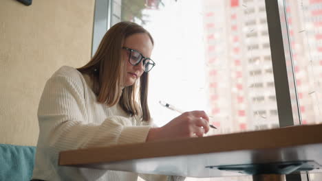 focused lady sketching on paper at wooden table near large window with soft daylight, urban view featuring tall residential building in background