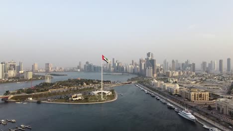 aerial view of the flag of the united arab emirates waving in the air city development in background, the national symbol of uae over sharjah's flag island