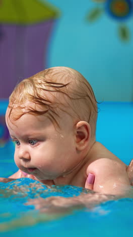 blond caucasian baby boy is held in water by woman. cute kid moves his hands in water making splashes. vertical video