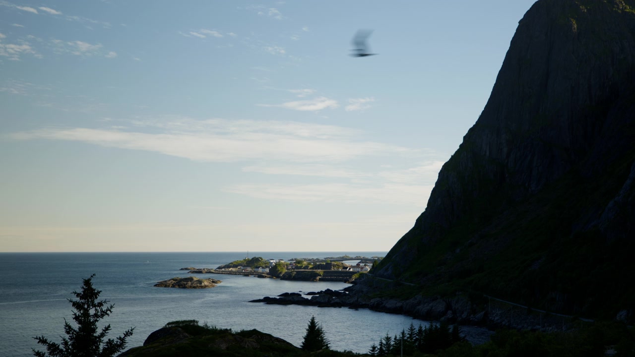 Høj mørk klippe rejser sig ved siden af en rolig havbugt med fjerne klippeøer og en lille kystbebyggelse i varmt eftermiddagslys