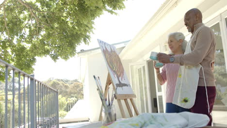 Happy-senior-biracial-couple-painting-and-drinking-coffee-at-balcony-at-home,-slow-motion