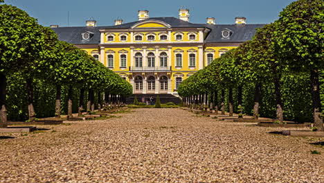 Timelapse-of-people-walking-across-gravel-path-of-green-trees-to-a-big-yellow-building