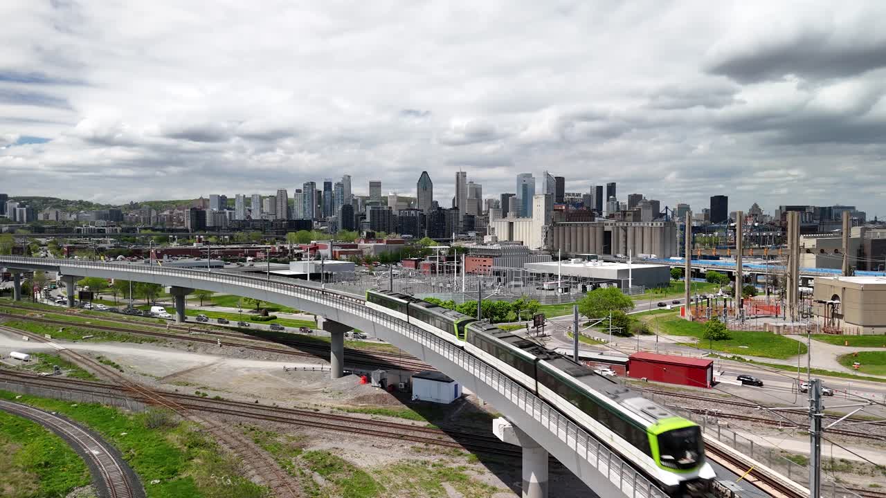 Aerial shot of Downtown Montreal skyline under overcast sky, features REM train moving towards camera and train tracks in urban setting.