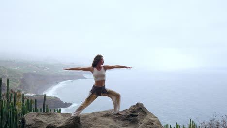 Equilibrándose-En-La-Pose-De-Guerrero-De-Yoga-Junto-Al-Océano,-La-Playa-Y-Las-Montañas-Rocosas,-La-Mujer-Encarna-Motivación,-Inspiración-Y-Dedicación-A-Un-Estilo-De-Vida-Saludable-Al-Aire-Libre.