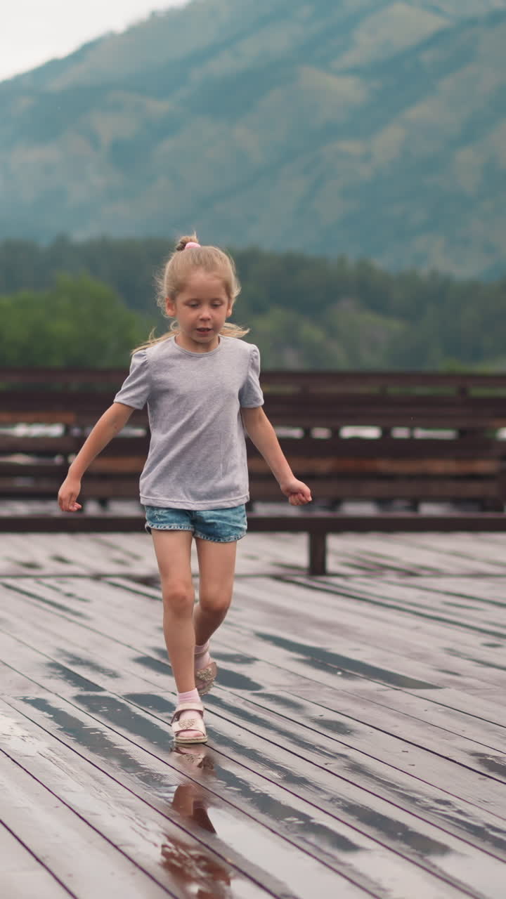 Playful little child girl in casual clothes runs along large empty wooden deck stepping small puddles at highland resort on spring day slow motion