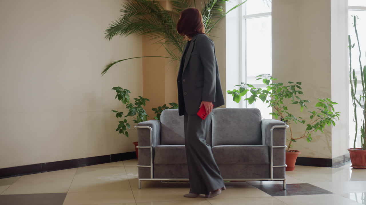Confident businesswoman in elegant gray suit standing beside sofa in stylish modern office lobby holding red smartphone surrounded by indoor plants natural sunlight professional setting