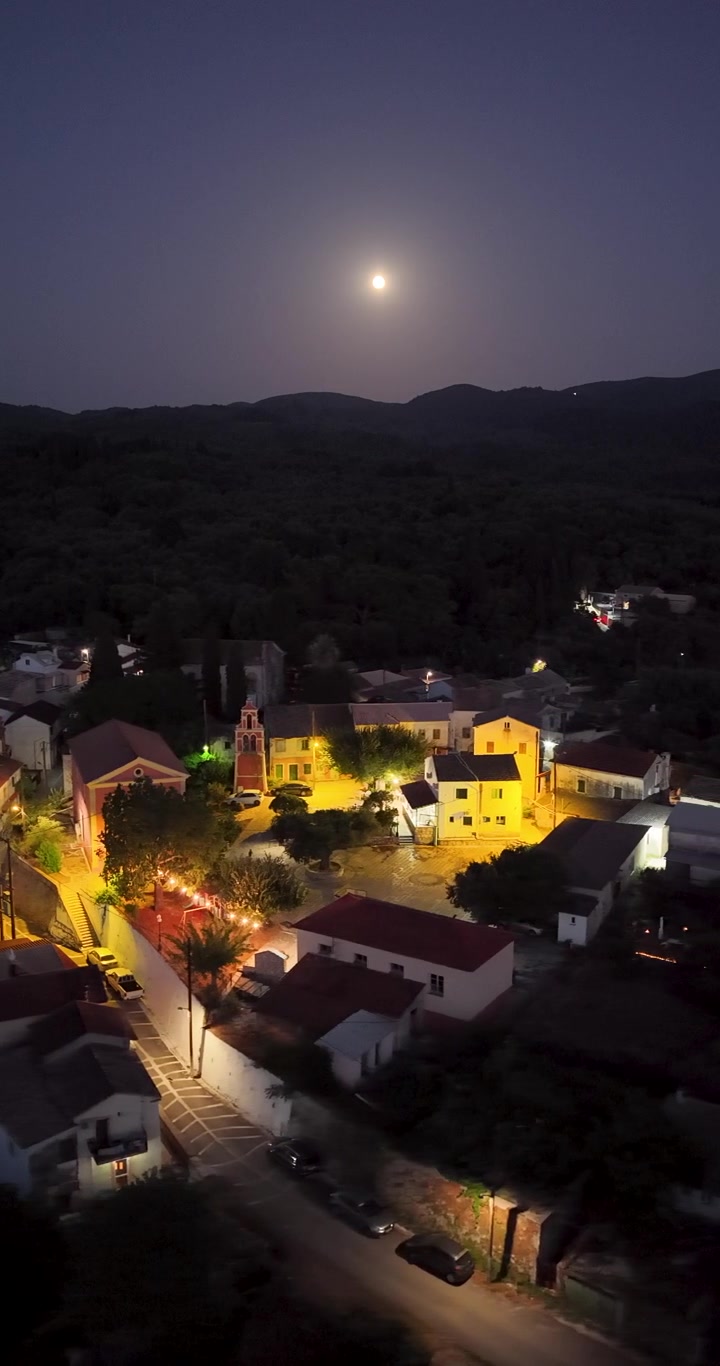 Filmagem aérea vertical de drone da vila de Agioi Douloi em Corfu, Grécia, à noite, exibindo praça e igreja brilhantes iluminadas sob uma noite de lua cheia.