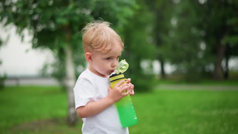 a little boy drinks from a bright green water bottle while outdoors, with a lush green background, his expression is focused as he holds the bottle