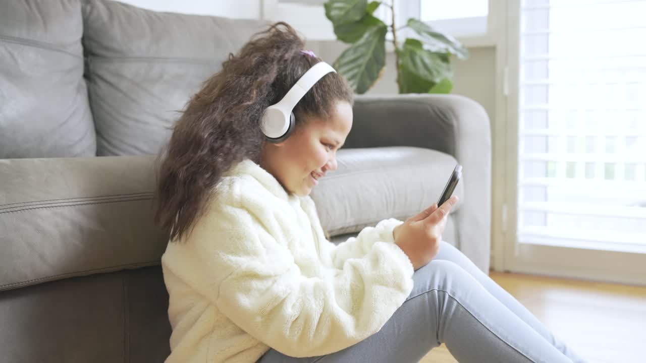 Ragazza preadolescente tiene in mano lo smartphone con le cuffie, appoggiata al divano e tocca lo schermo in soggiorno.