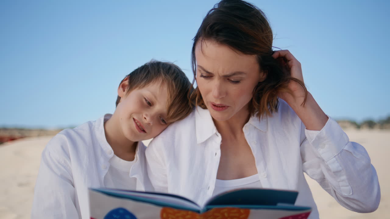 Spensierata famiglia che legge un libro seduta vicina su una spiaggia sabbiosa, primo piano su una giovane madre.