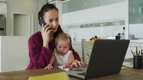 caucasian mother holding her baby talking on smartphone and using laptop while working from home