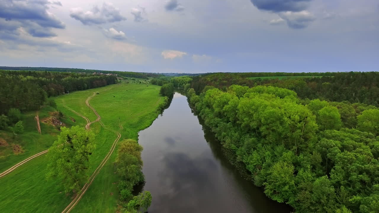 Narrow river with meadow on one side and forest on another. Cloudy sky reflecting in the water. Top view.