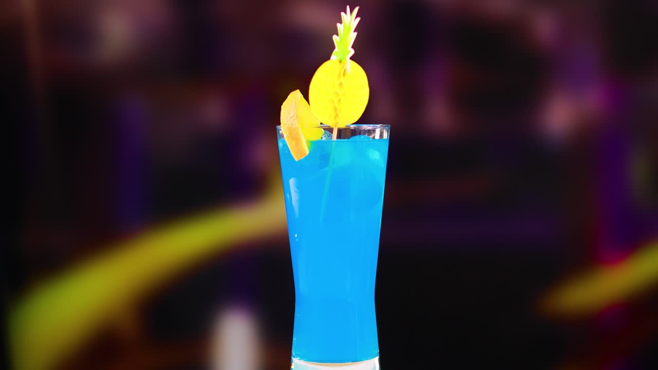 A vibrant blue cocktail is garnished with fruit and mint, set against a blurred background, highlighting its refreshing appeal
