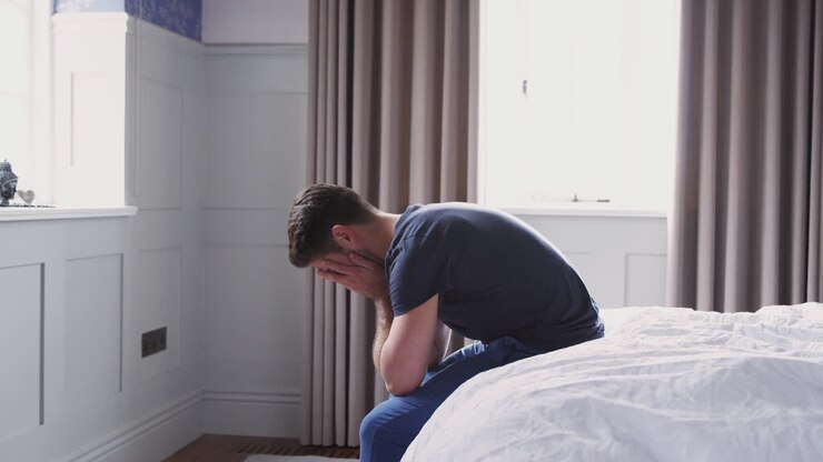 Man Wearing Pajamas Suffering With Depression Sitting On Bed At Home