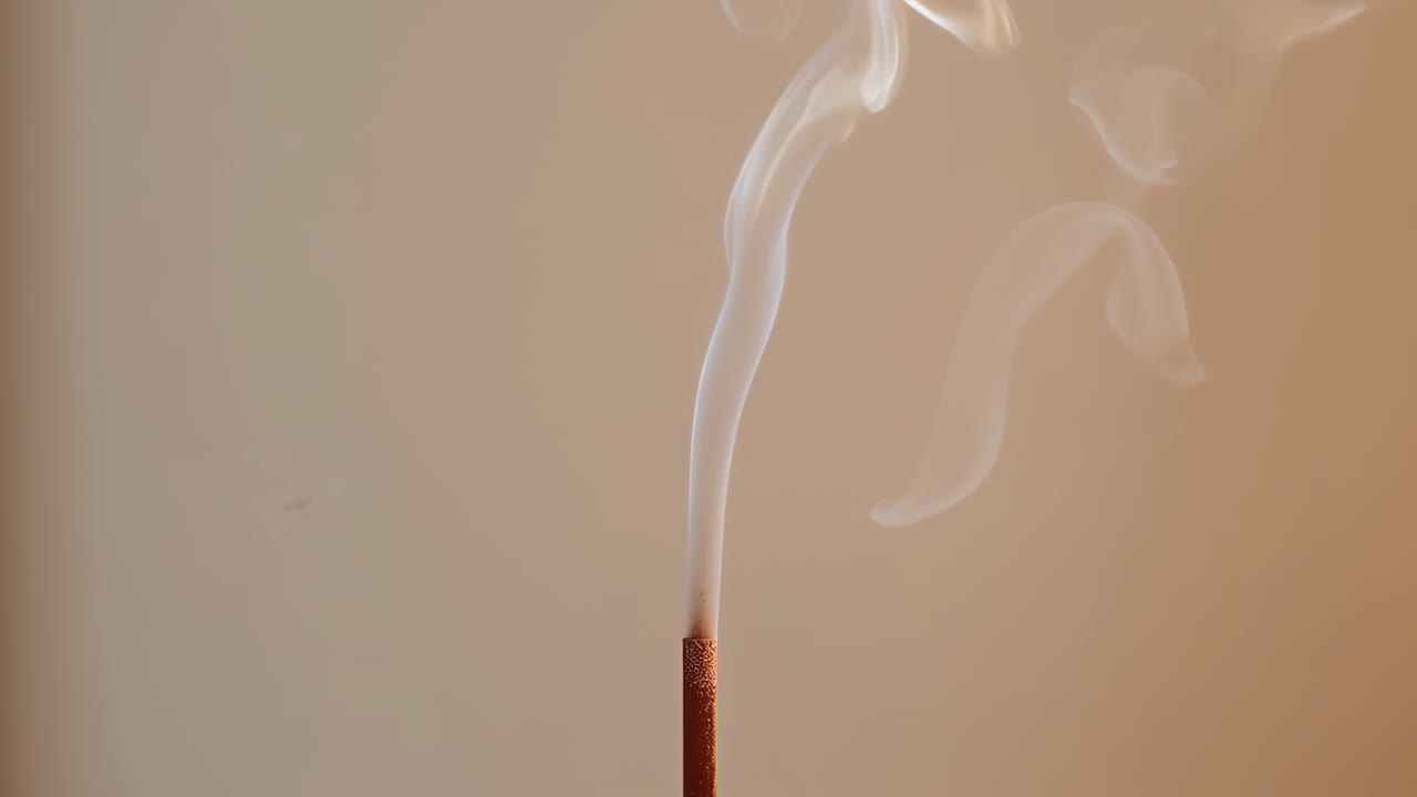 Bâtonnet d’encens fin incandescent dégageant de la fumée vers le haut dans une maison, montrant des cendres et un mur beige chaud