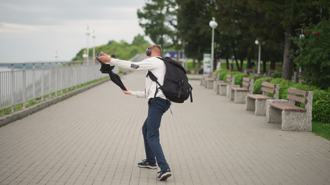 흰색 옷을 입은 남자가 물가의 산책로에서 배낭을 메고 장난스럽게 우산을 흔들고 있어요. 하늘은 흐리고 가랑비가 내리고, 벤치와 난간 옆에서 활기찬 걸음걸이와 과장된 포즈를 취하고 있으며, 자연스러운 도시의 여유로운 순간을 담고 있습니다.