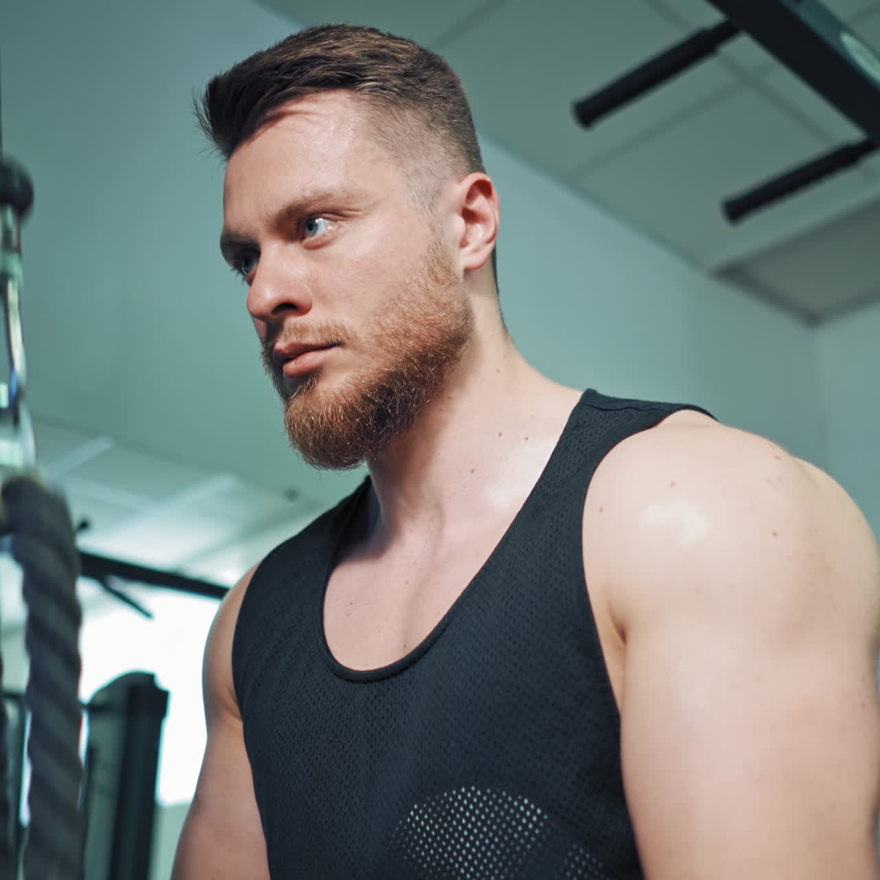 Concentrated athlete training his arm biceps in sports club. Portrait of young sportsman doing his workout on a modern simulator by pulling rope.