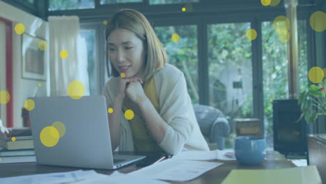 animation of yellow spots floating over asian woman having a video call on laptop at home