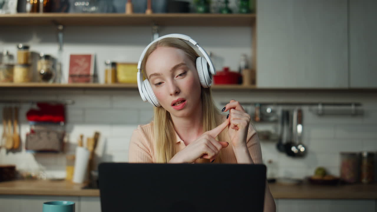 Woman teacher lecturing online sitting at kitchen with laptop wearing headphones