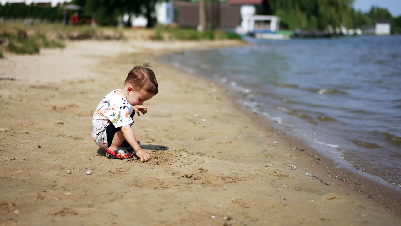 Lovely dark-haired toddler sits on the sandy shore. Kid touches wet sand and looks at water.