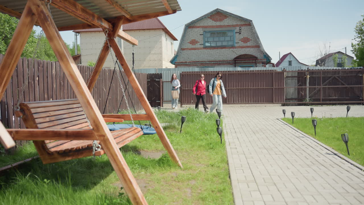 Three siblings stroll toward recreational center, passing wooden swing under bright sky, cobblestone path, green grass, rustic fence, quaint houses craft serene outdoor scene captured in sunny moment