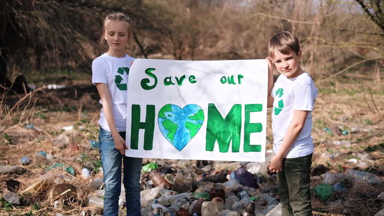Kirlenmiş bir alanda plastik çöp toplama etkinliğinde ekolojiyi savunan poster tutan oğlan ve kız çocuk, tişörtlerinde geri dönüşüm işaretleri. Ağır çekim