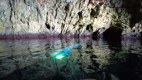 Kayaker-observing-a-mysterious-scene-with-a-beam-of-light-shimmering-in-the-water-inside-a-sea-cave,-Vis-island,-Adriatic-Sea,-Croatia