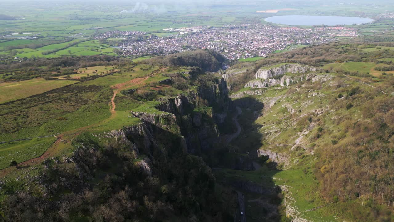 Aerial forward shot showcasing Cheddar Gorge cliffs, fields, village, and lake.
