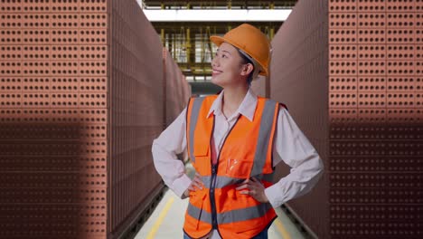 asian female engineer wearing safety helmet looking around while standing with arms akimbo with red brick packed in stacks are stored