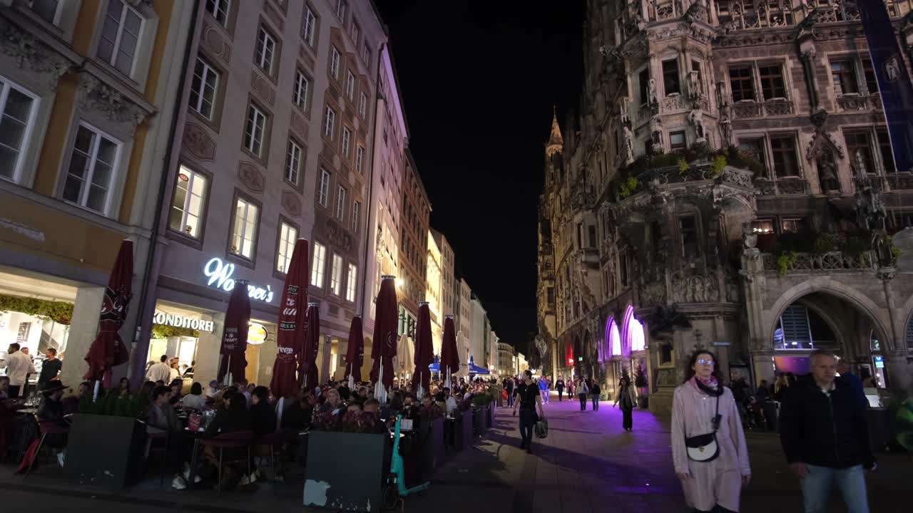 A wide-angle night view of Munich, capturing people shopping and sightseeing beneath an illuminated building.
