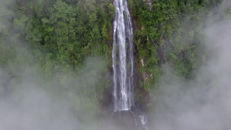 Aerial-tilt-down-of-clouds-over-Las-Lajas-misty-waterfall-streaming-down-rocky-pond-surrounded-by-rainforest,-San-Luis-Morete,-Costa-Rica